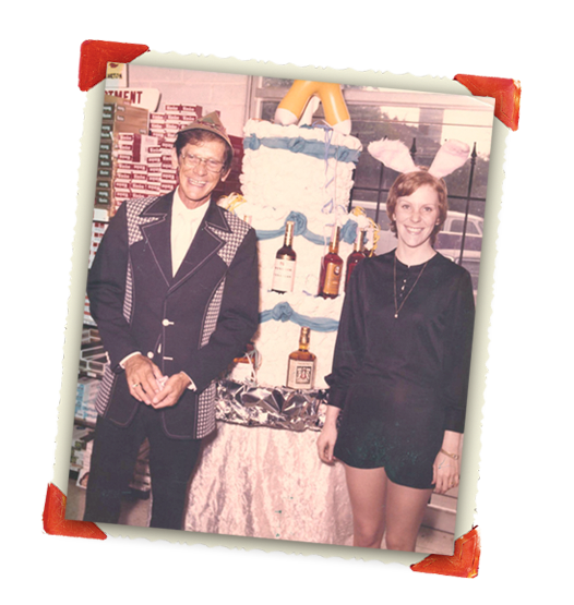 Vintage photograph of two people standing next to a birthday cake decorated with liquor bottles